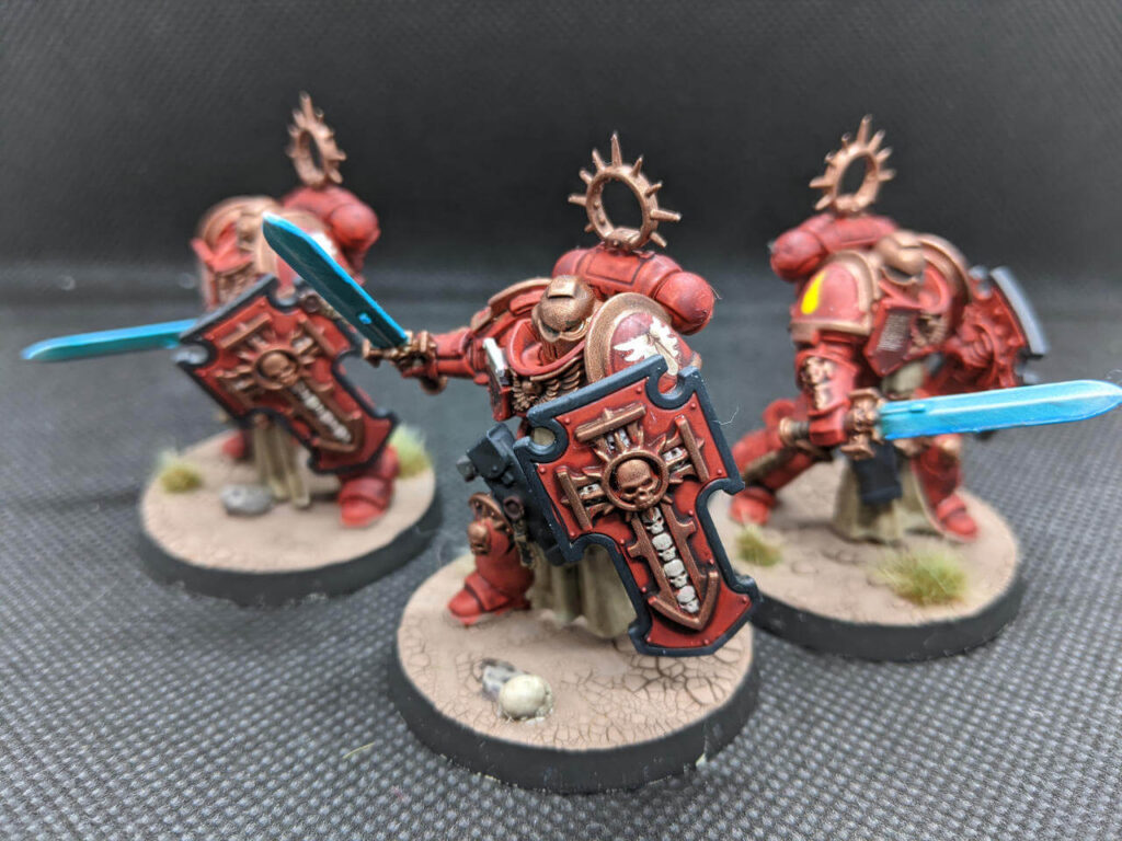 A close-up view of three Warhammer 40k miniatures depicting Blood Angels Bladeguard Veterans. The models are wearing red power armor with intricate detailing, including large red shields adorned with skull emblems and winged insignias. Each figure holds a bright blue energy sword, and the base features a rocky terrain with patches of dry grass.