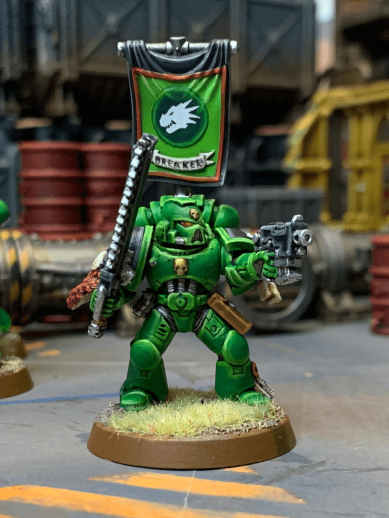 A painted model of a Salamanders Space Marine, holding a chainsword and banner with a dragon emblem.